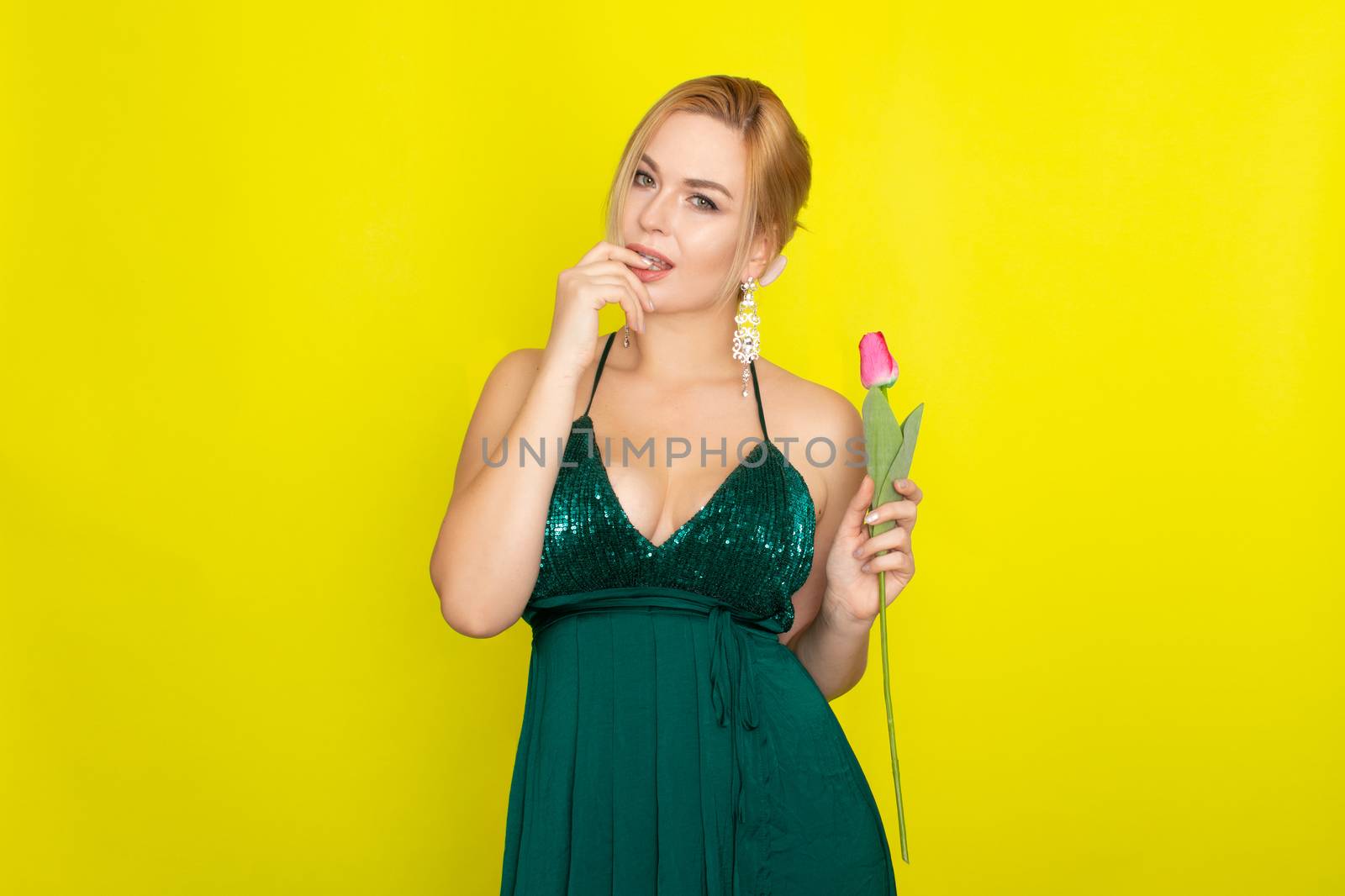 Blonde woman in green evening dress holding one tulip in her hands over yellow background