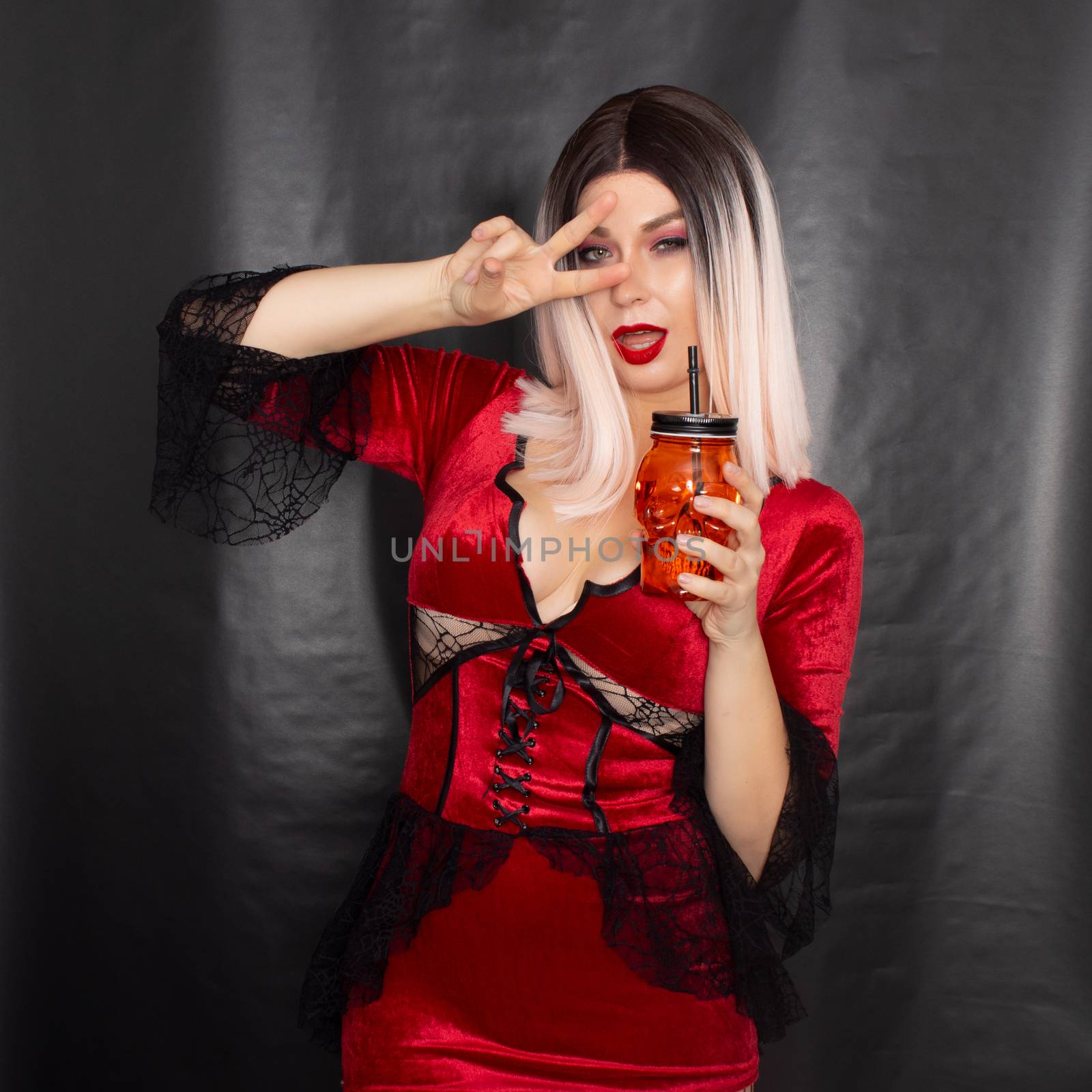 Young beautiful blonde woman in a red vampire dress holds an orange glass in the shape of a skull in her hands