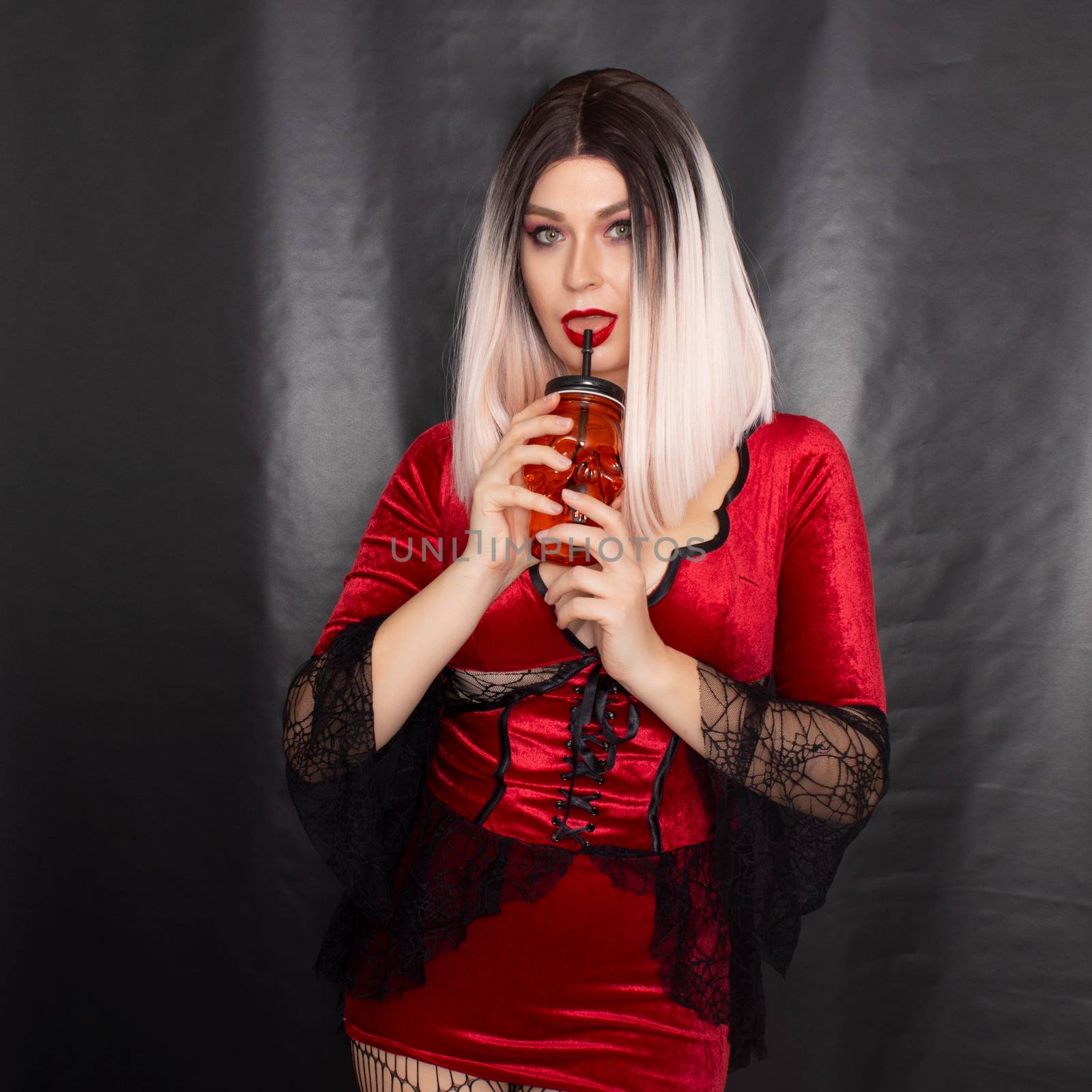 Young beautiful blonde woman in a red vampire dress holds an orange glass in the shape of a skull in her hands