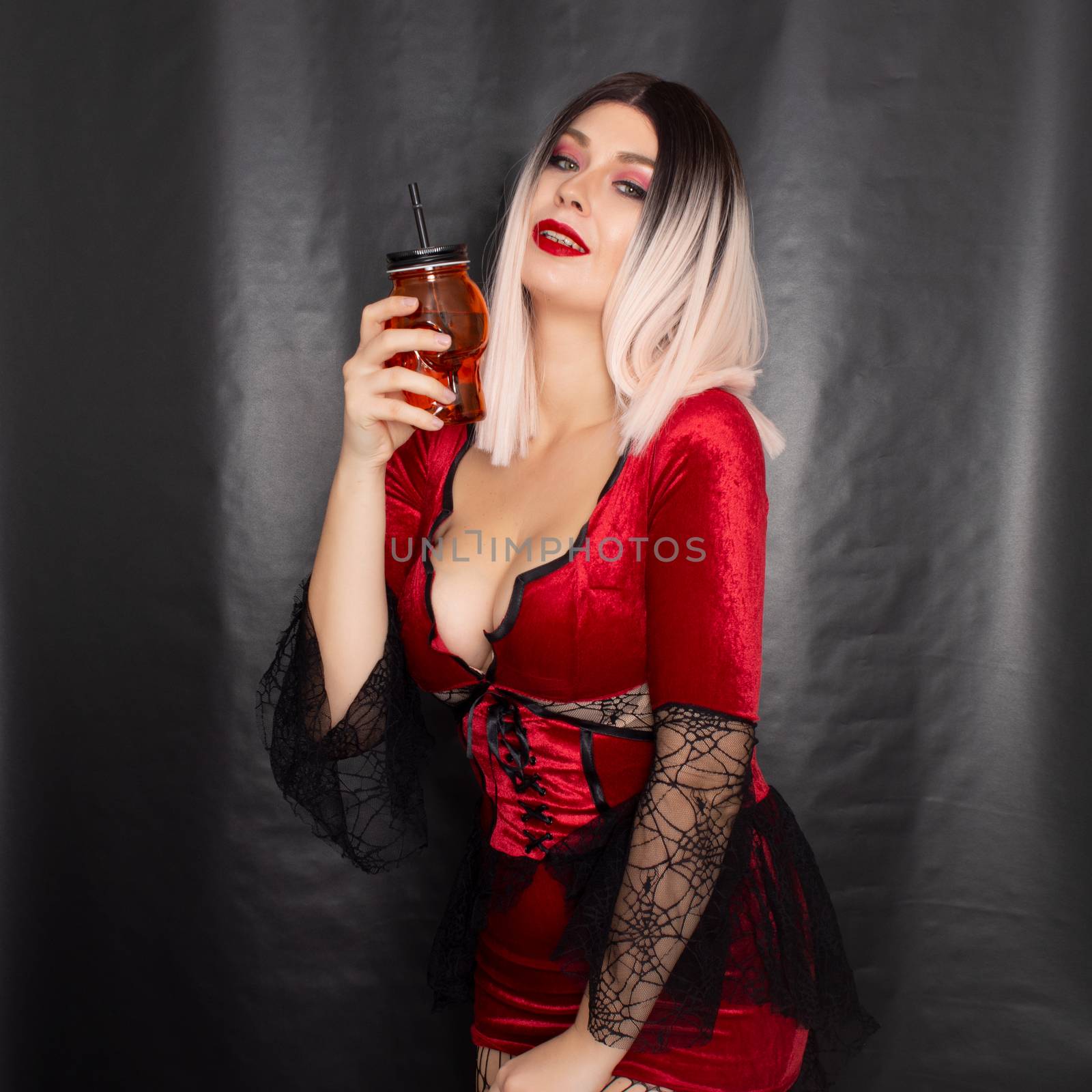 Young beautiful blonde woman in a red vampire dress holds an orange glass in the shape of a skull in her hands