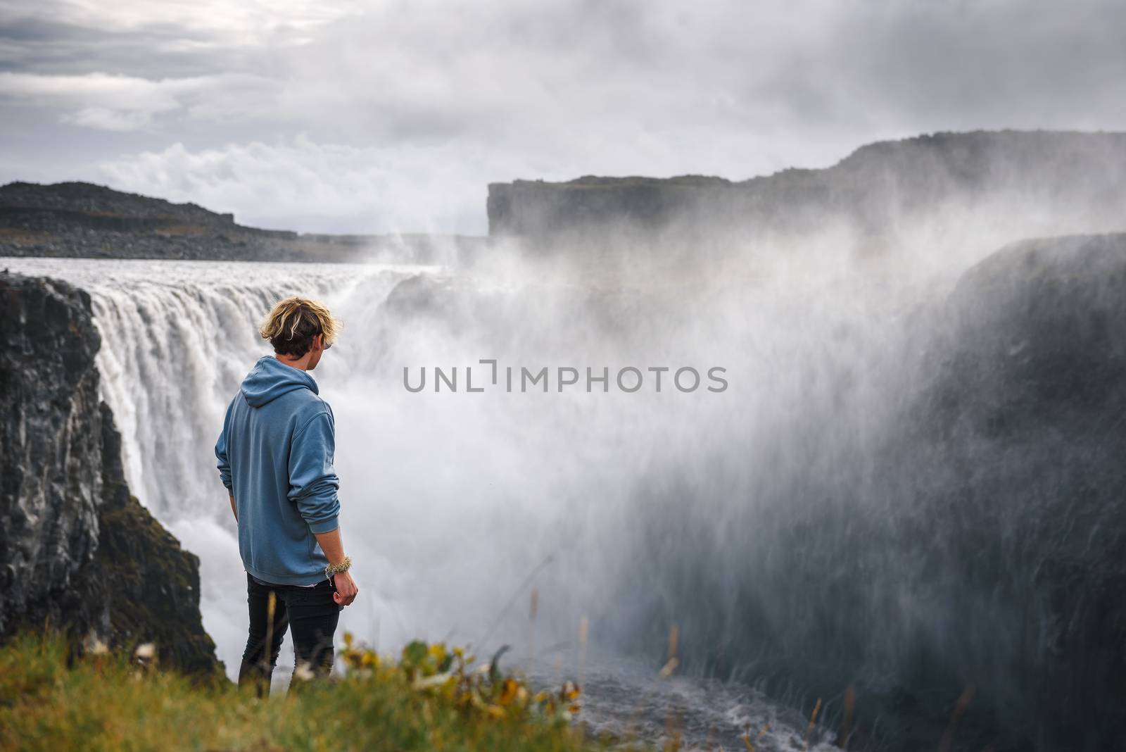 Young hiker standing at the edge of the Dettifoss waterfall located on the Jokulsa a Fjollum river in Iceland. Dettifoss is the second most powerful waterfall in Europe after the Rhine Falls.