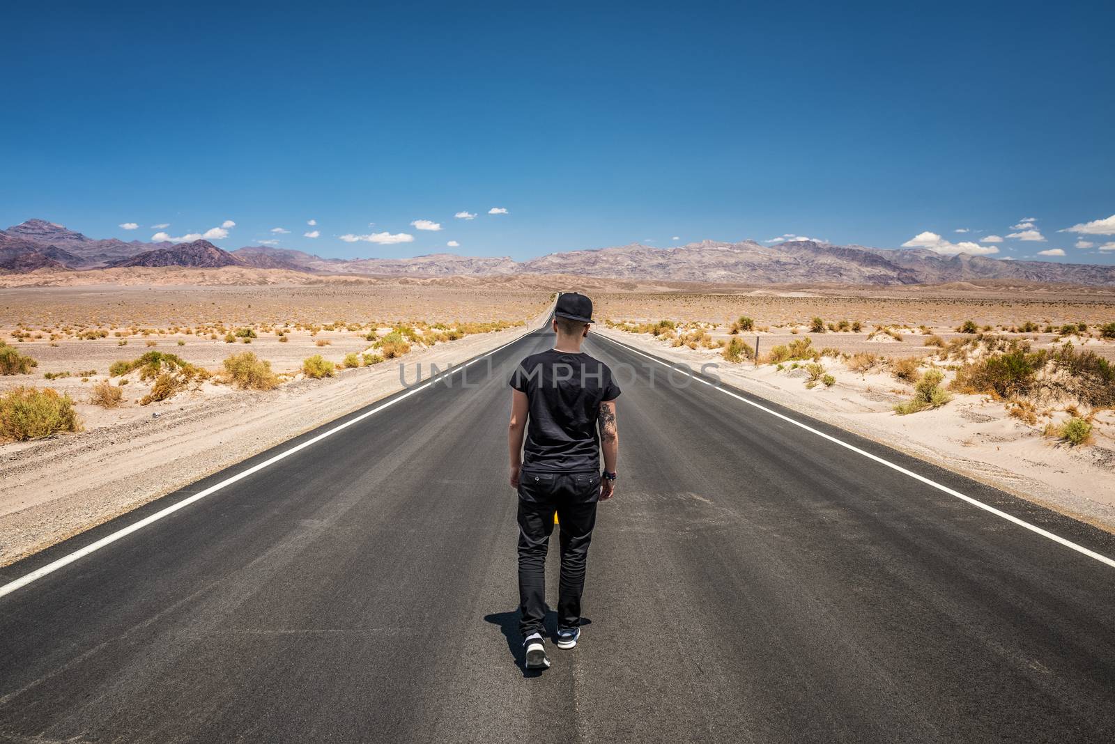 Young man walking alone through an empty street in the desert of Death Valley National Park on a bright sunny day. Loneliness, sadness , depression and solitude concept in nature.