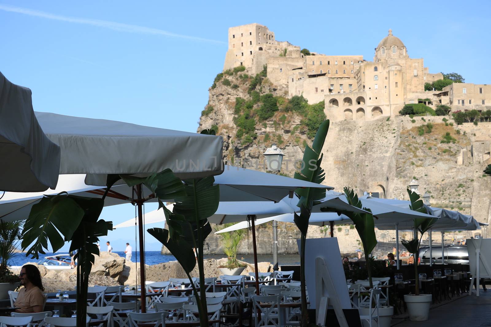 Ischia, Naples, Italy, About the July 2019. Umbrellas and tables of a romantic seaside restaurant. In the background the Aragonese Castle of Ischia.