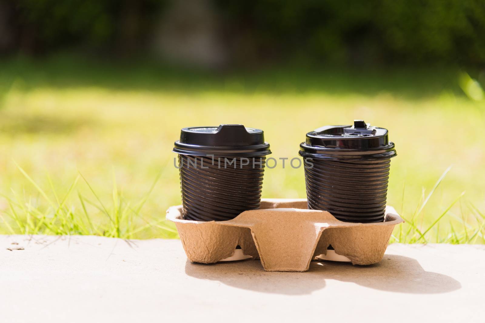 Close up of two black paper cups hot coffee to takeaway on the floor outside the cafe on the garden place of green leaves color background for copy space with sunlight in the morning day