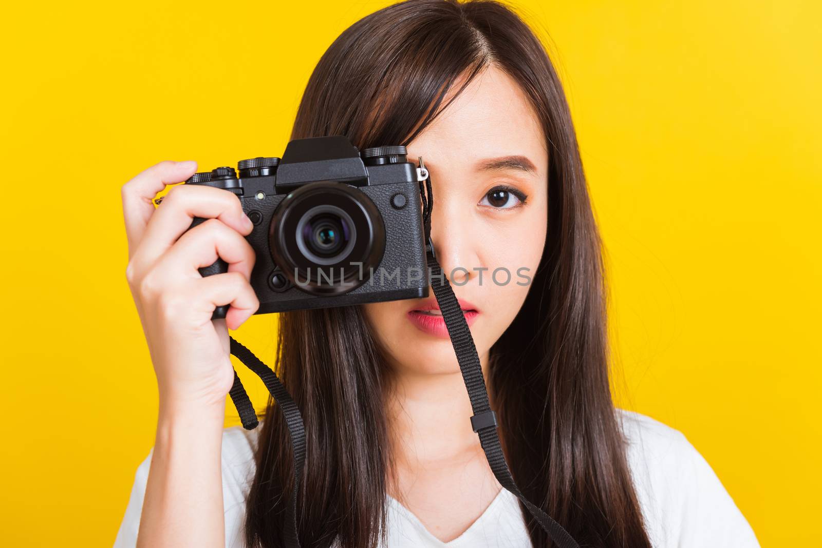 Portrait of happy Asian beautiful young woman photographer taking a picture and looking viewfinder on retro digital mirrorless photo camera ready to shoot, studio shot isolated on yellow background