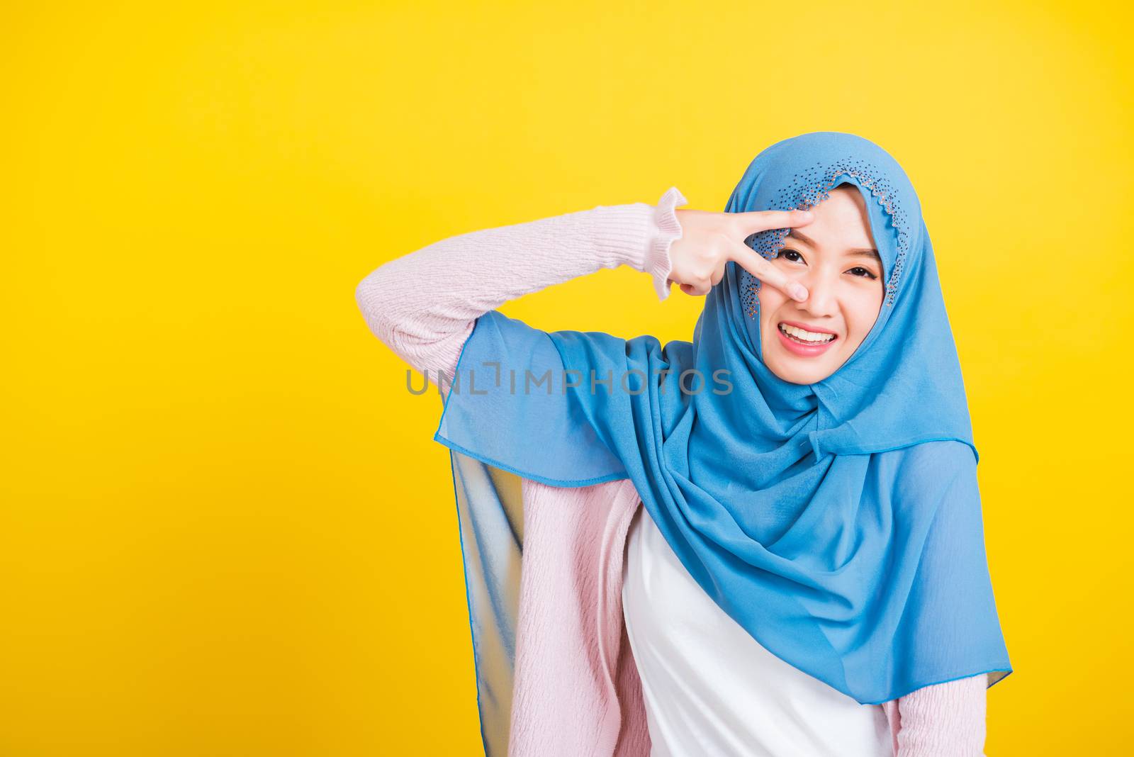 Asian Muslim Arab, Portrait of happy beautiful young woman Islam religious wear veil hijab smile she showing gesture hand two finger V-sign symbol near eyes isolated yellow background