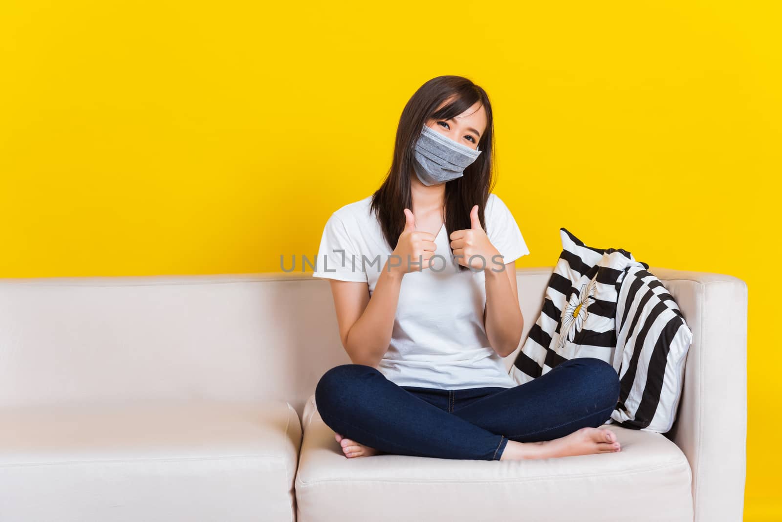 Portrait Asian of beautiful young woman sitting on sofa wearing medical face mask protective show finger thumb up for good sign during Coronavirus studio shot isolated on yellow background