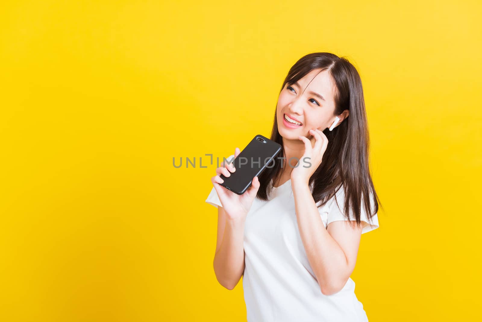 Portrait Asian of a happy beautiful young woman holding a mobile smart phone and wearing wireless headphones listening to music from smartphone studio shot isolated on yellow background