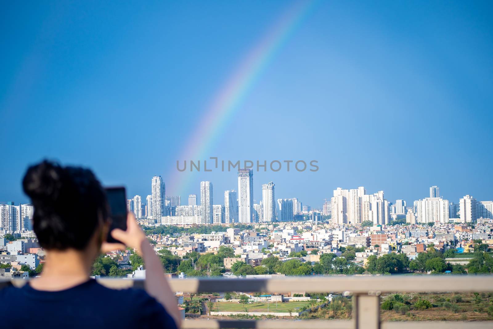 Young indian girl wearing hair in a bun and with blue dress photographing rainbow over gurgaon delhi noida cityscape on a monsoon day by Shalinimathur