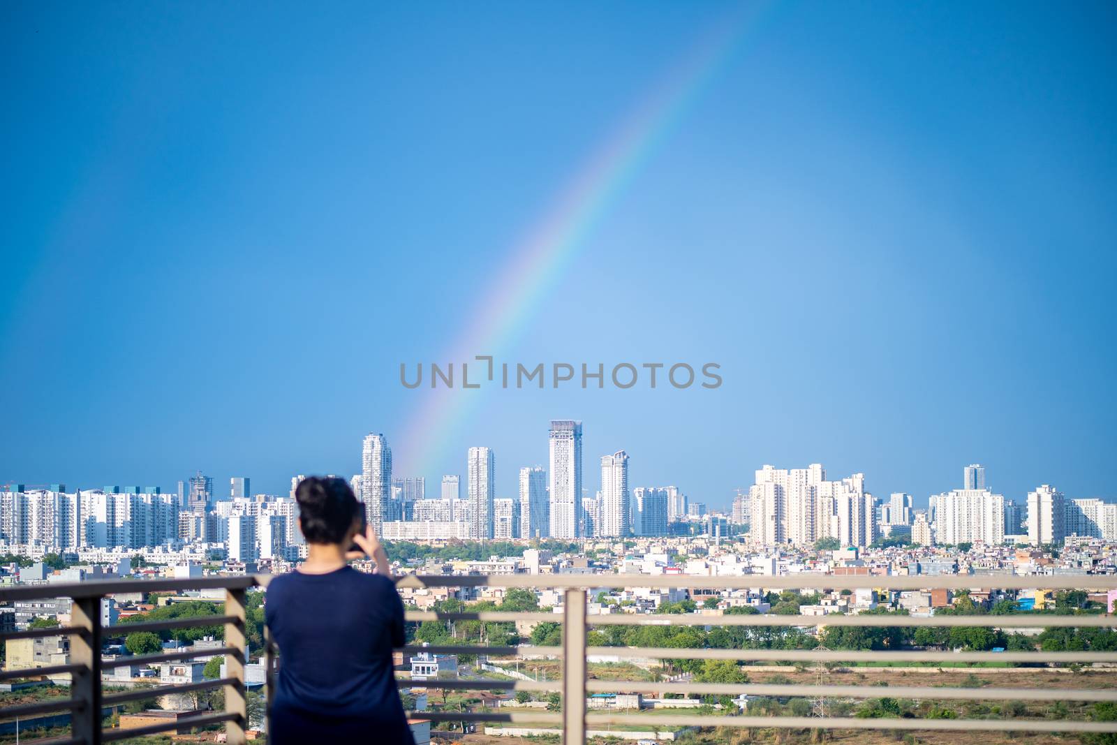 Young indian girl wearing hair in a bun and with blue dress photographing rainbow over gurgaon delhi noida cityscape on a monsoon day. Photo of the natural phenomenon that took place in the indian capital some months back