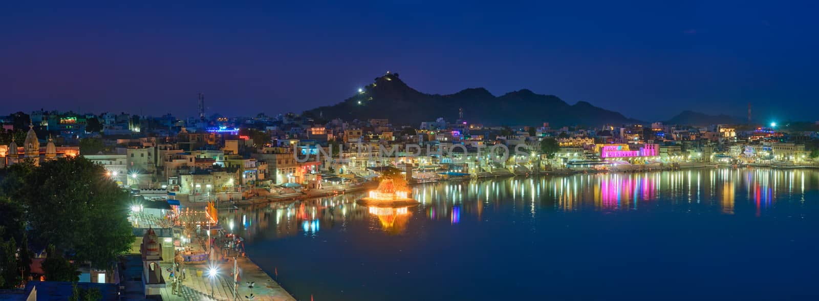 View of famous indian hinduism pilgrimage town sacred holy hindu religious city Pushkar with Brahma temple, aarti ceremony, lake and ghats illuminated at sunset. Rajasthan, India. Horizontal pan