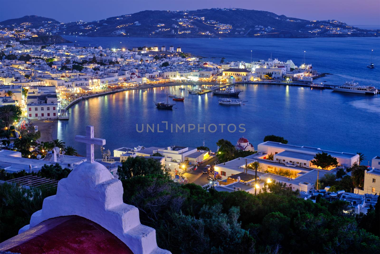 Mykonos Chora town Greek tourist holiday vacation destination with famous windmills port with boats yachts illuminated in evening blue hour with St. Basil church cross. Mykonos, Cyclades islands, Greece