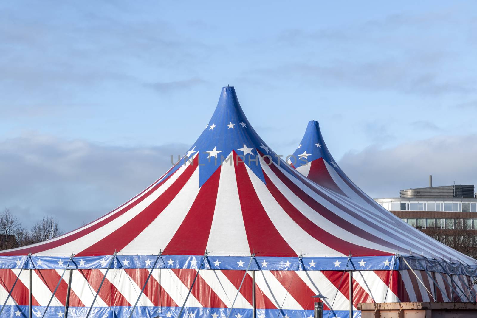 Red and white circus tent topped with bleu starred cover against a sunny blue sky with clouds by ankorlight