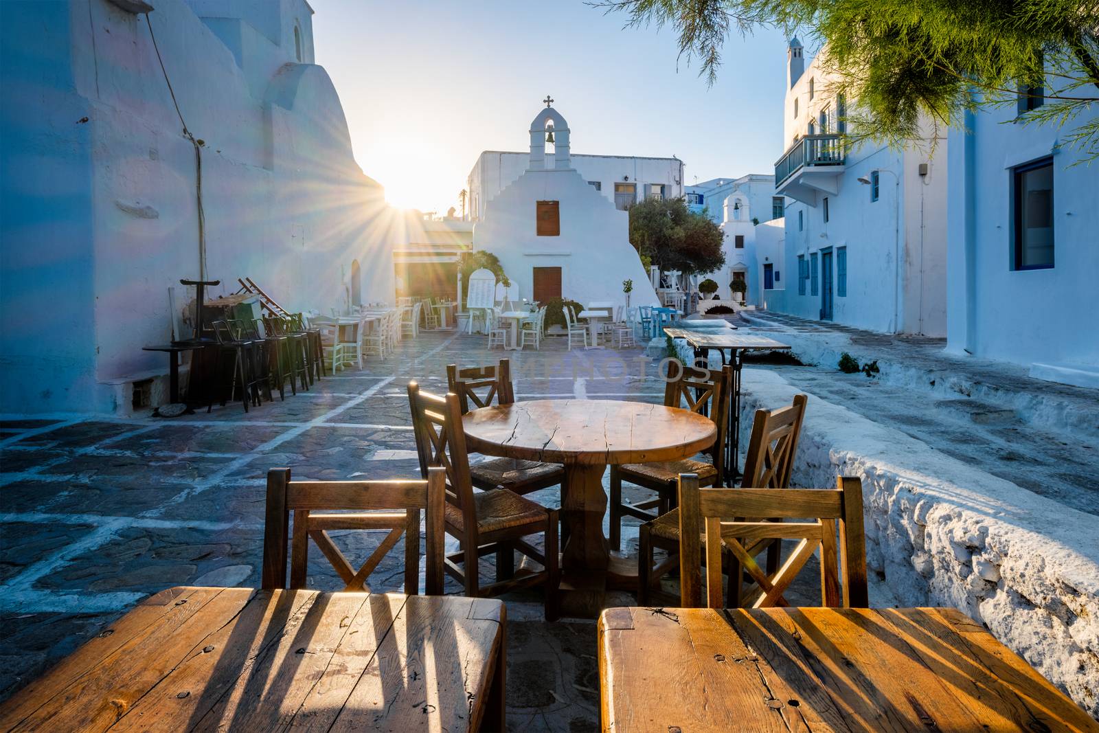 Tourist Greece scene - restaurant cafe table in picturesque scenic narrow streets with traditional whitewashed houses blue doors windows Chora town in famous tourist attraction Mykonos island, Greece