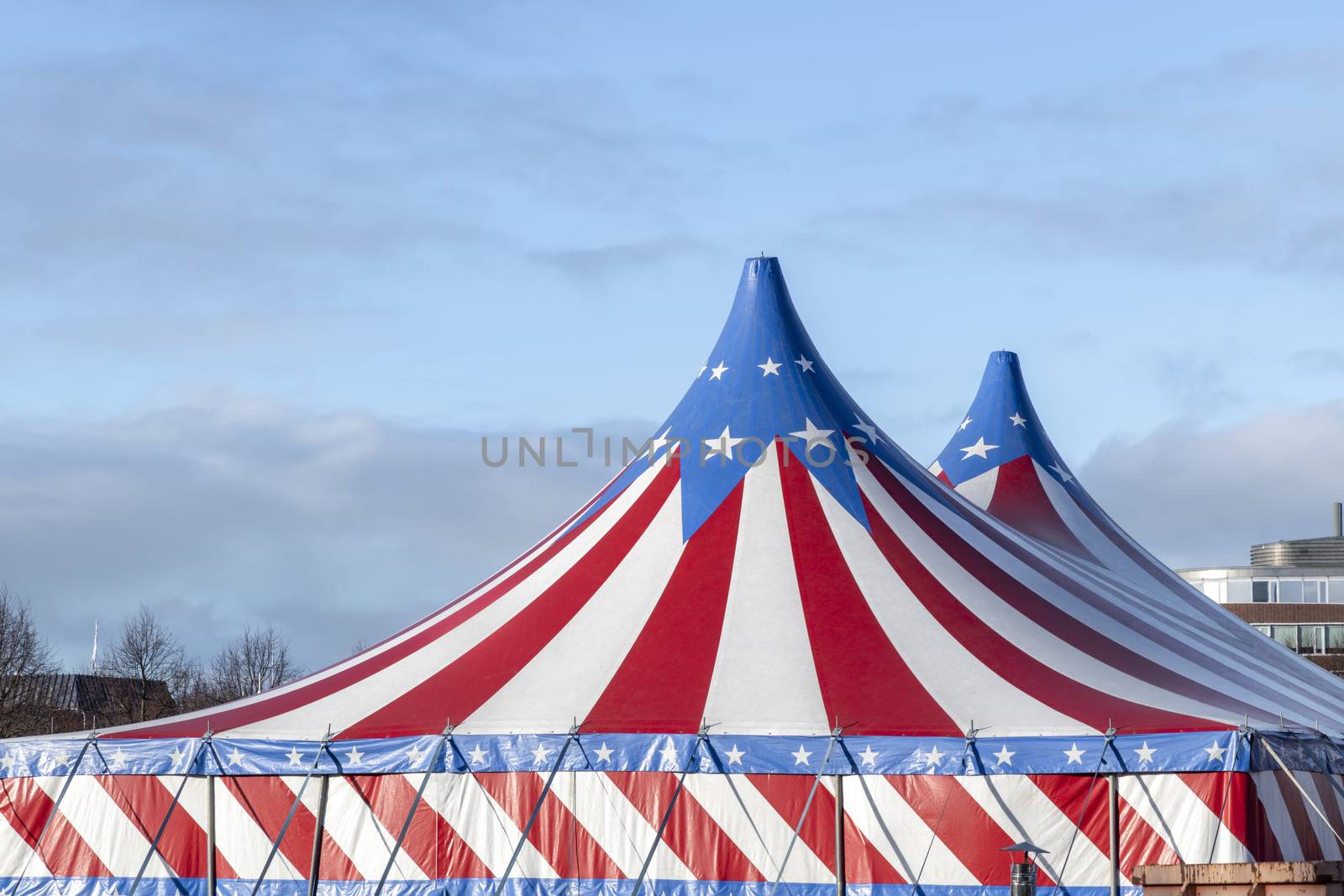 Red and white circus tent topped with bleu starred cover against a sunny blue sky with clouds by ankorlight