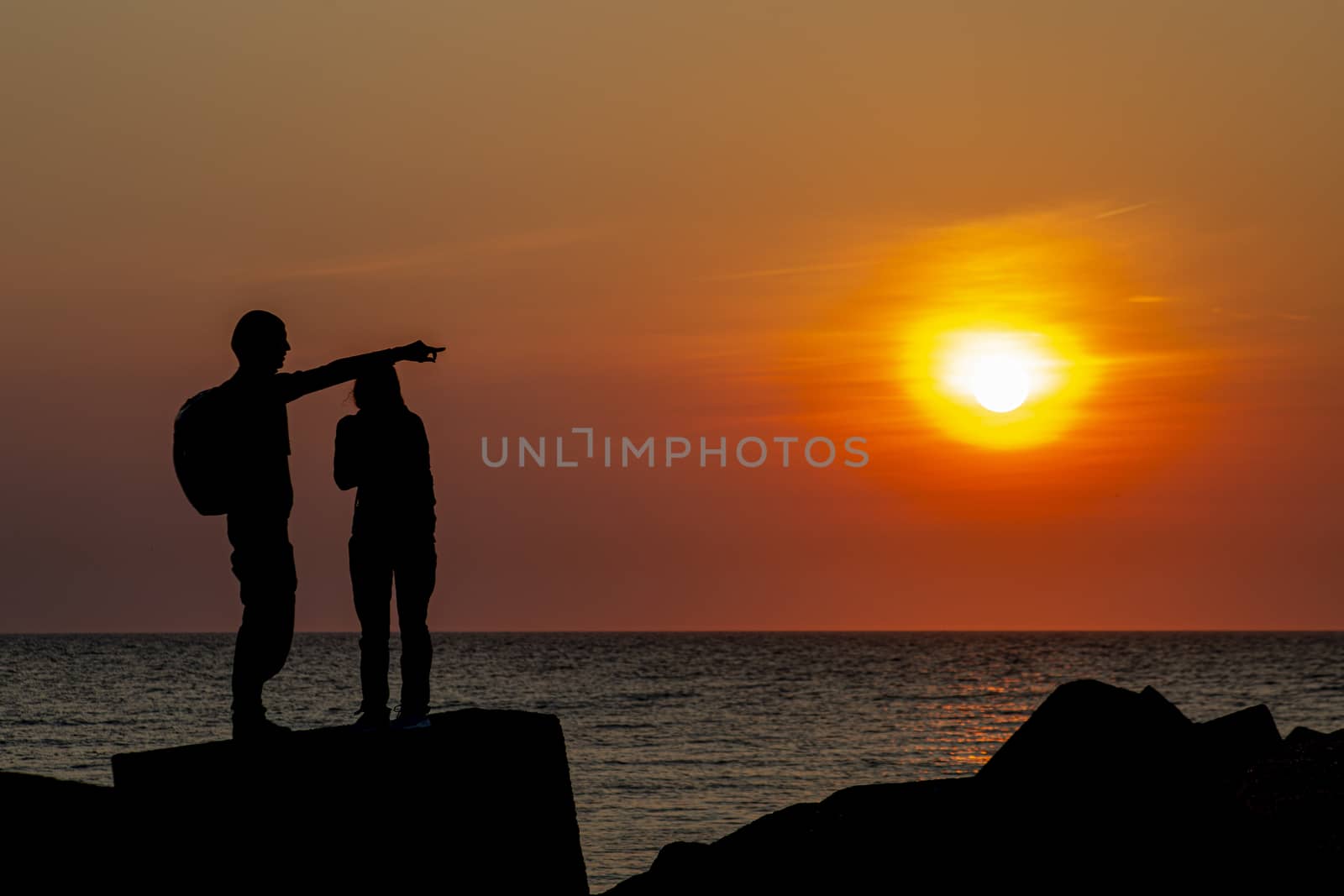 Silhouette of a young couple standing on the concrete pier protection blocks at Scheveningen under a vivid sunset moment, The Hague, Netherlands