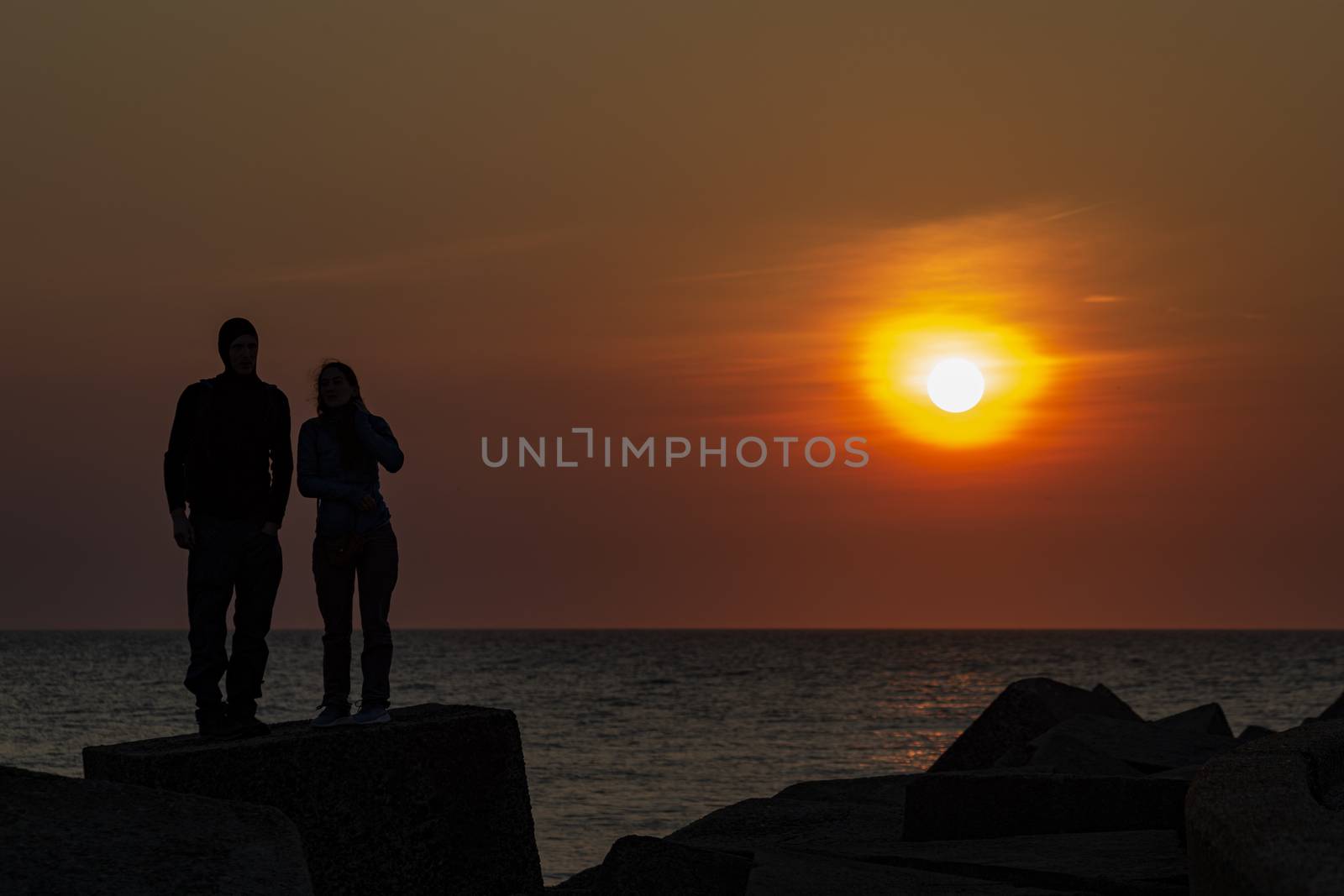 Silhouette of a young couple standing on the concrete pier protection blocks at Scheveningen under a vivid sunset moment, The Hague, Netherlands