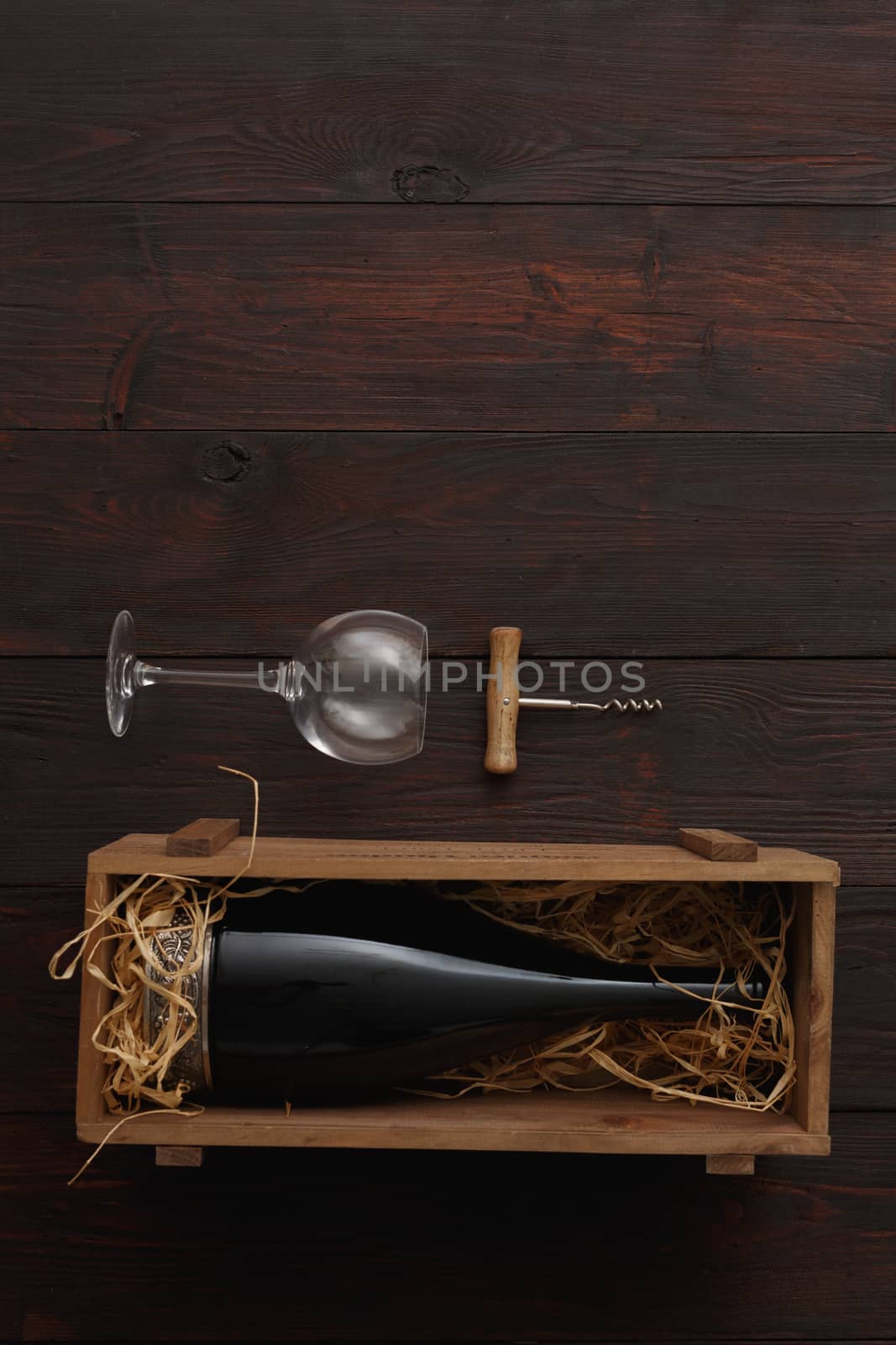 Bottle of red wine in wooden crate filled with straw next to to empty glasses with corkscrew on wooden table. Flat lay, above view, copy space,Chiaroscuro styled