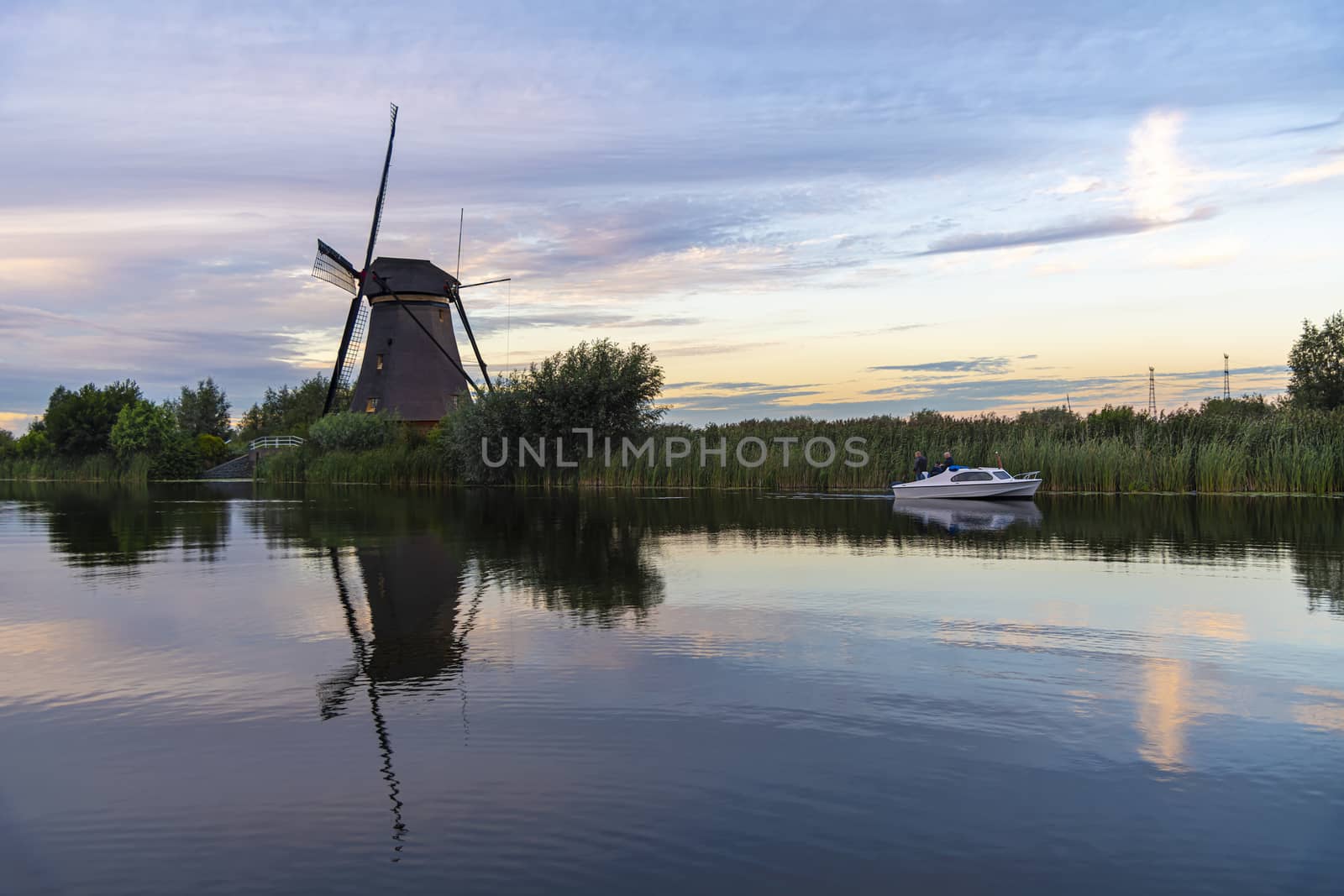 Dutch windmill laying along the canal with wild grass blown by strong winds at the early sunset moment