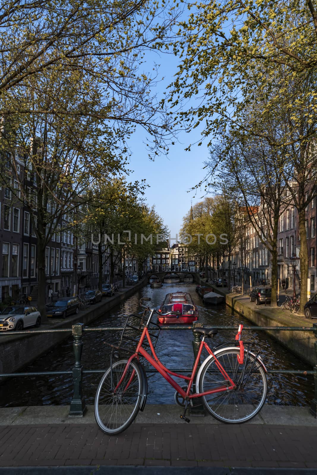 Red bicycle parking on the bridge over the calm water of a canal with a boat giving a round trip to tourists, Amsterdam, Netherlands by ankorlight