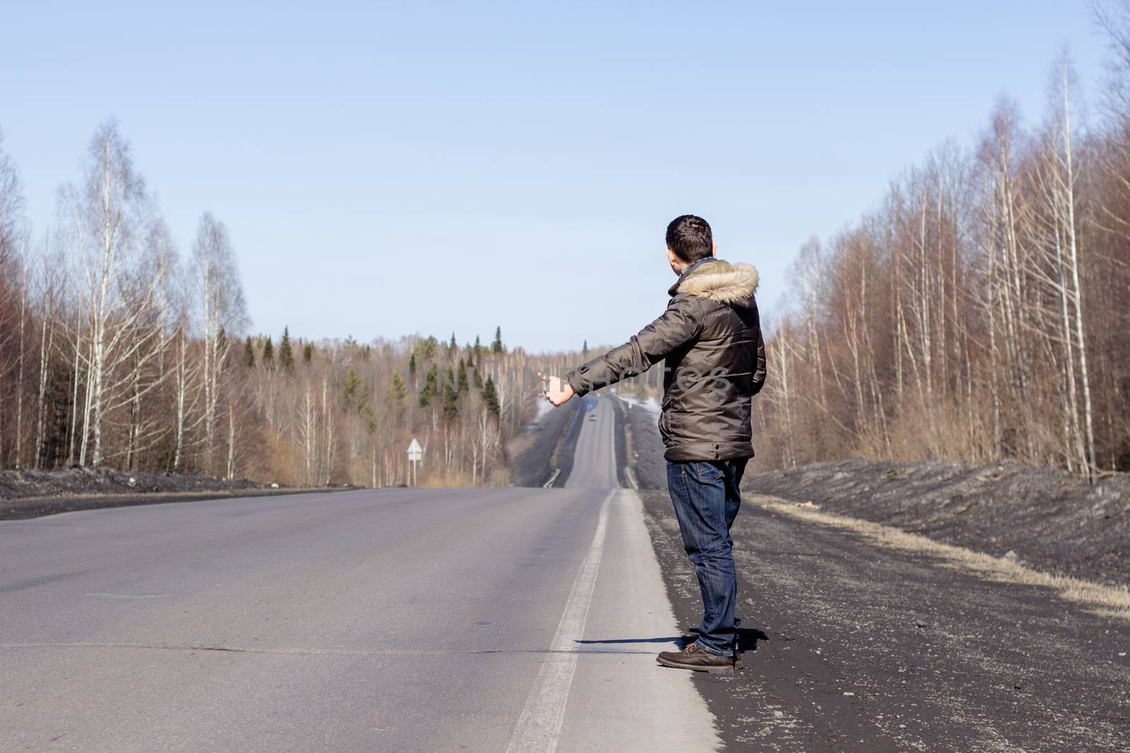 A young guy is walking on the side of the road, in a jacket. Road in the woods in early spring..