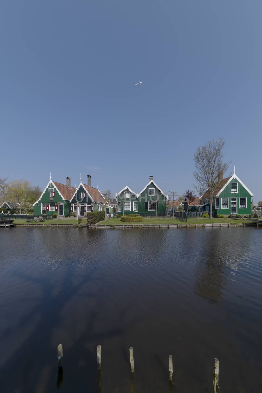 Plane flying above the Dutch green rural houses at the edge of the canals in the clear blue sunny sky by ankorlight
