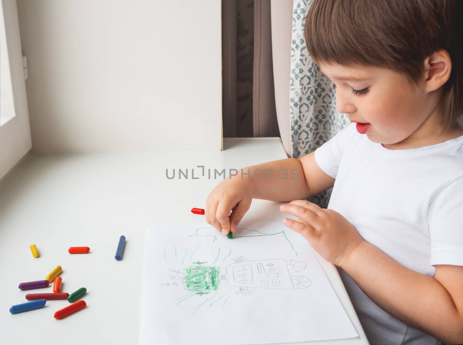 Toddler draws funny robot. Kid uses wax crayons. Smiling boy paints on paper with pencils on white windowsill.