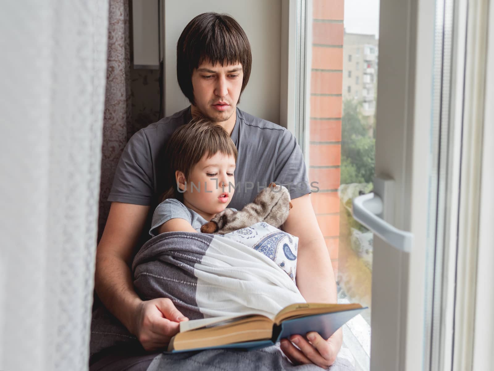 Father reads book to his son. Cozy family time on windowsill while raining outside. Toddler boy sits together with fluffy toy cover himself with a blanket.