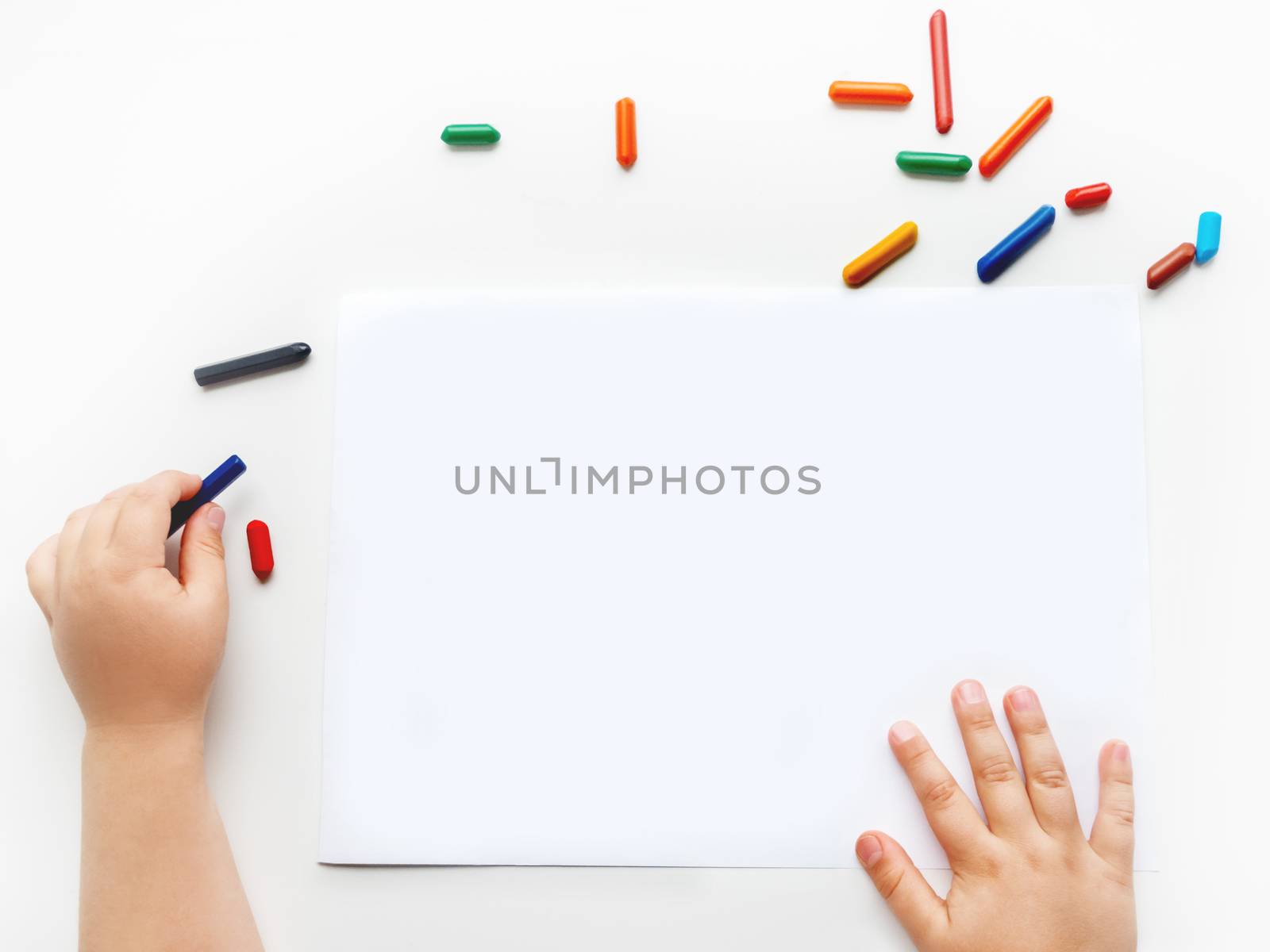 Left-handed toddler prepare to draw something on clear white sheet of paper. Kid uses wax crayons. Top view on child's hands and pencils on white background.