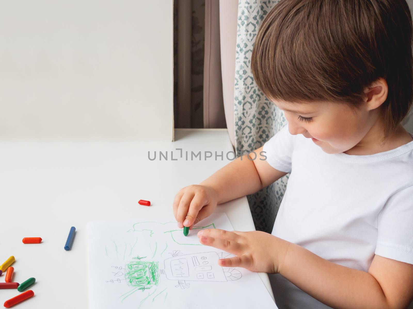 Toddler draws funny robot. Kid uses wax crayons. Smiling boy paints on paper with pencils on white windowsill.