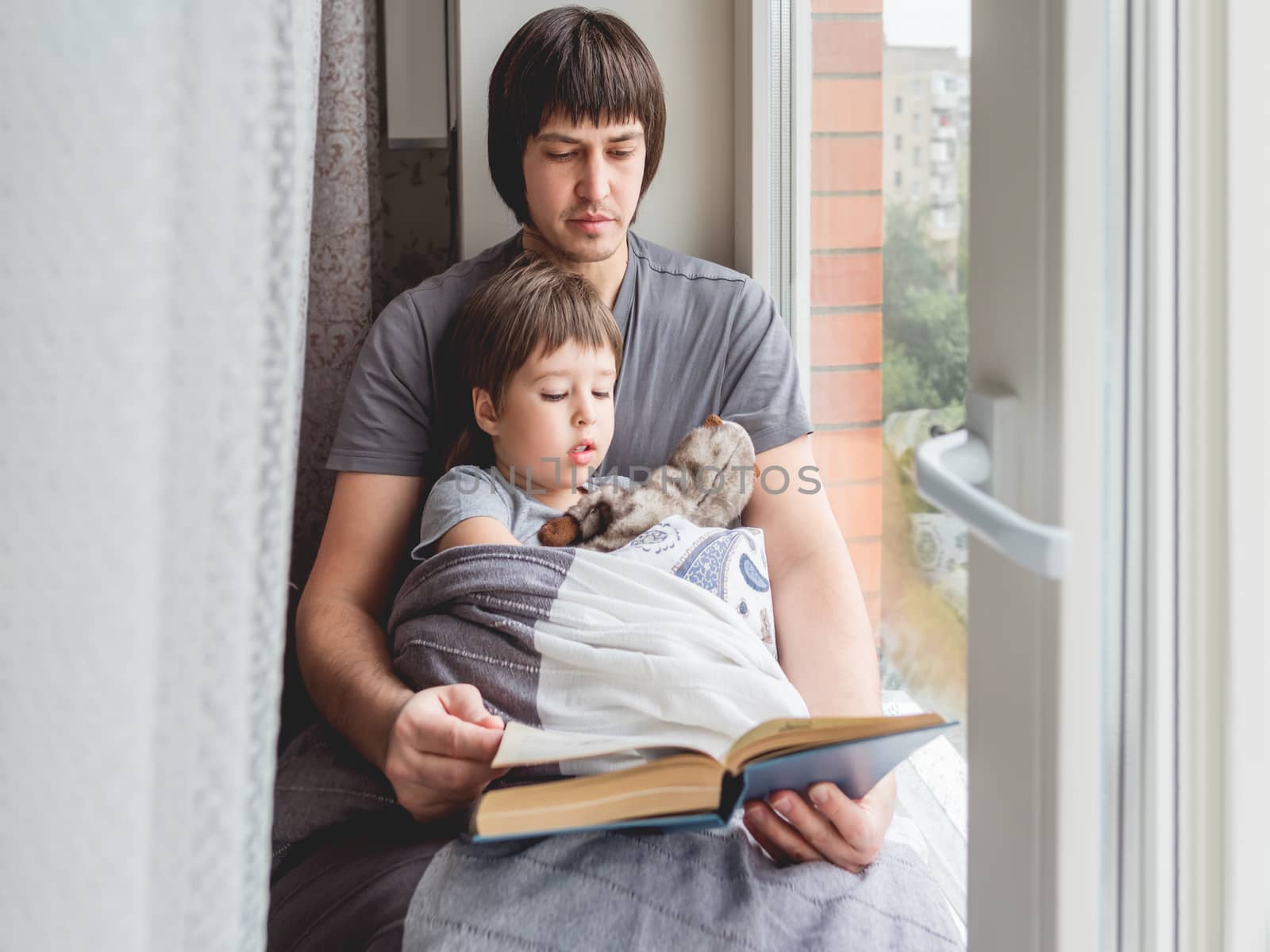 Father reads book to his son. Cozy family time on windowsill whi by aksenovko