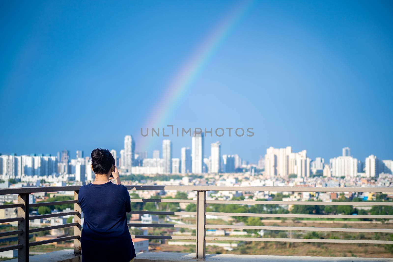 Young indian girl wearing hair in a bun and with blue dress photographing rainbow over gurgaon delhi noida cityscape on a monsoon day by Shalinimathur