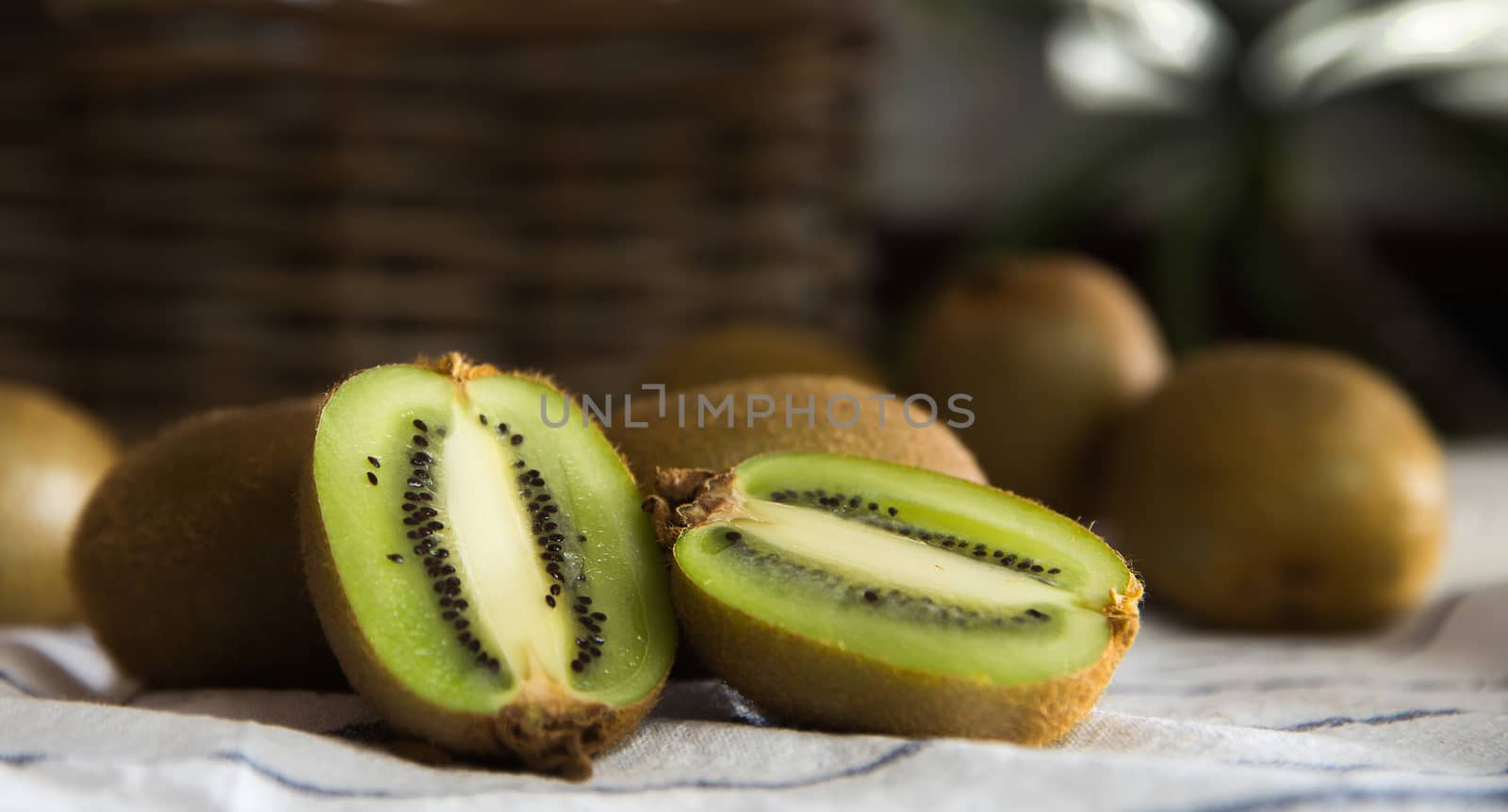 Close-up of a group of kiwis, one of them cut in half. Dark background with a wicker basket. On a white tablecloth with blue lines.