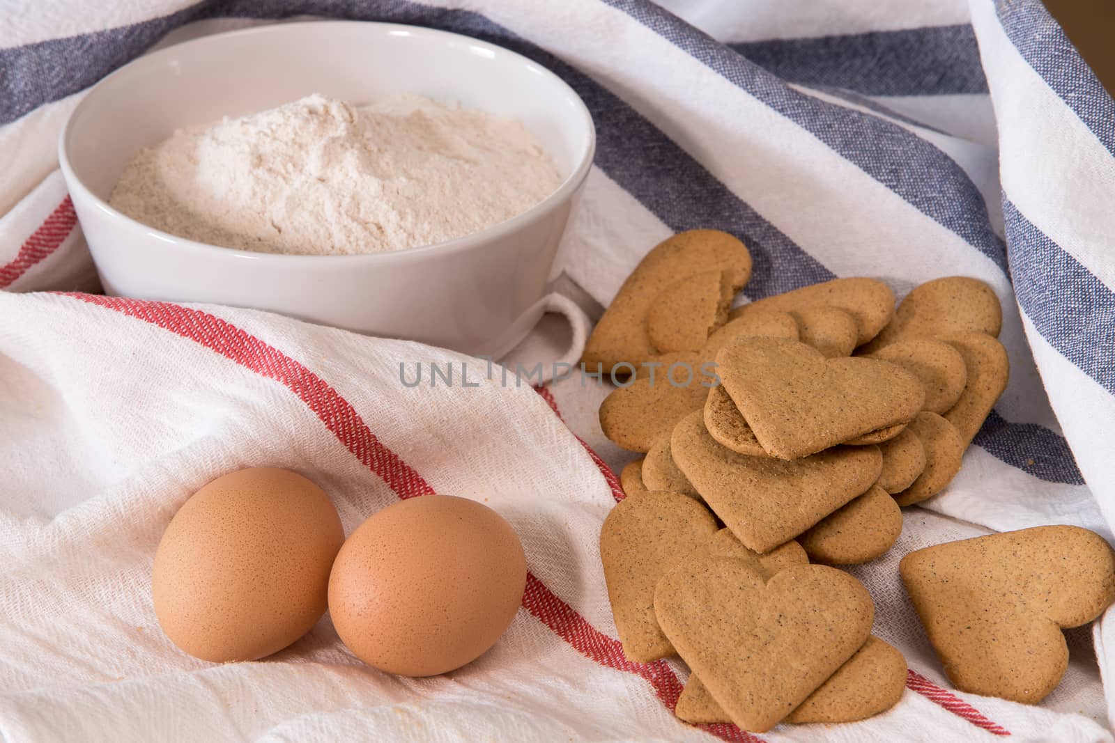 Gourmet close up with a bowl full of flour, heart shape cookies, and two eggs in white kitchen cloths with red and blue border, on a wooden board