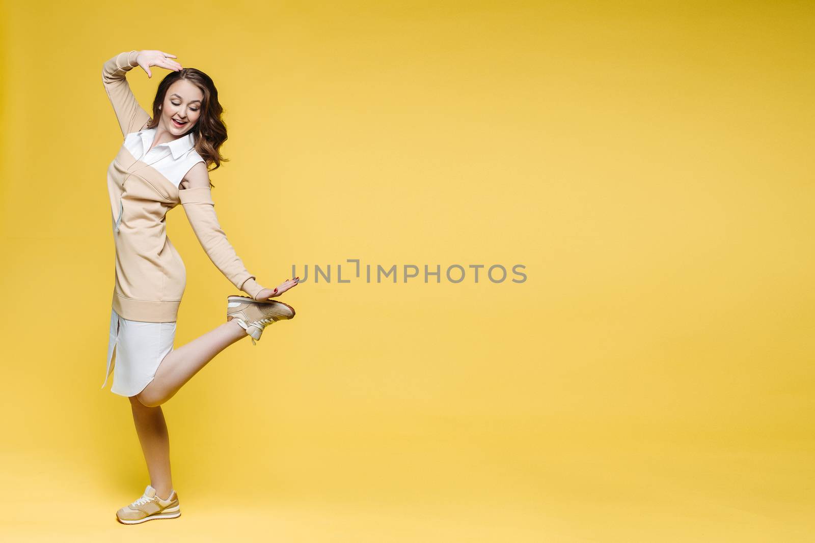 Positivity and fashionable brunette woman happy posing, smiling, holding leg and touching hair, looking down. Stylish girl with long wavy hair jumping, dancing at yellow studio. Concept of fashion.