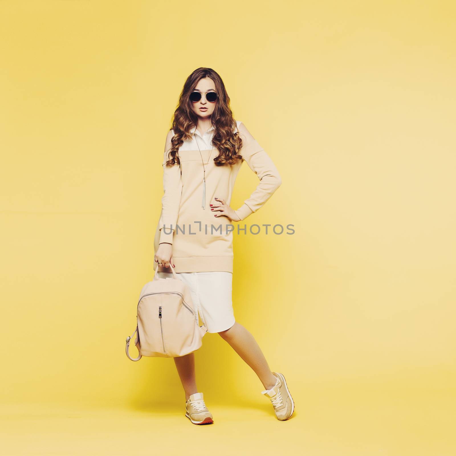 Fashionable beautiful brunette girl with wavy hair, perfect make up after beauty salon, wearing in white shirt and beige sweatshirt posing with hands on waist. Woman after shopping. Yellow background.