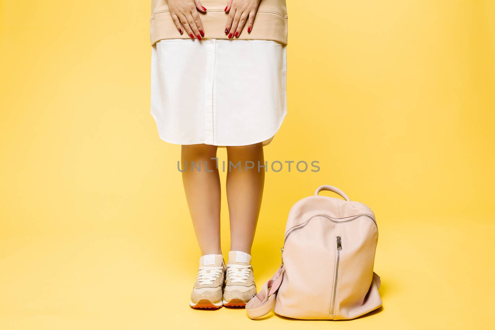 Crop of stylish incognito woman posing at studio in fashionable beige dress with elements of white, standing near pink leather bag on floor.Swag look of hipster woman against yellow studio background.