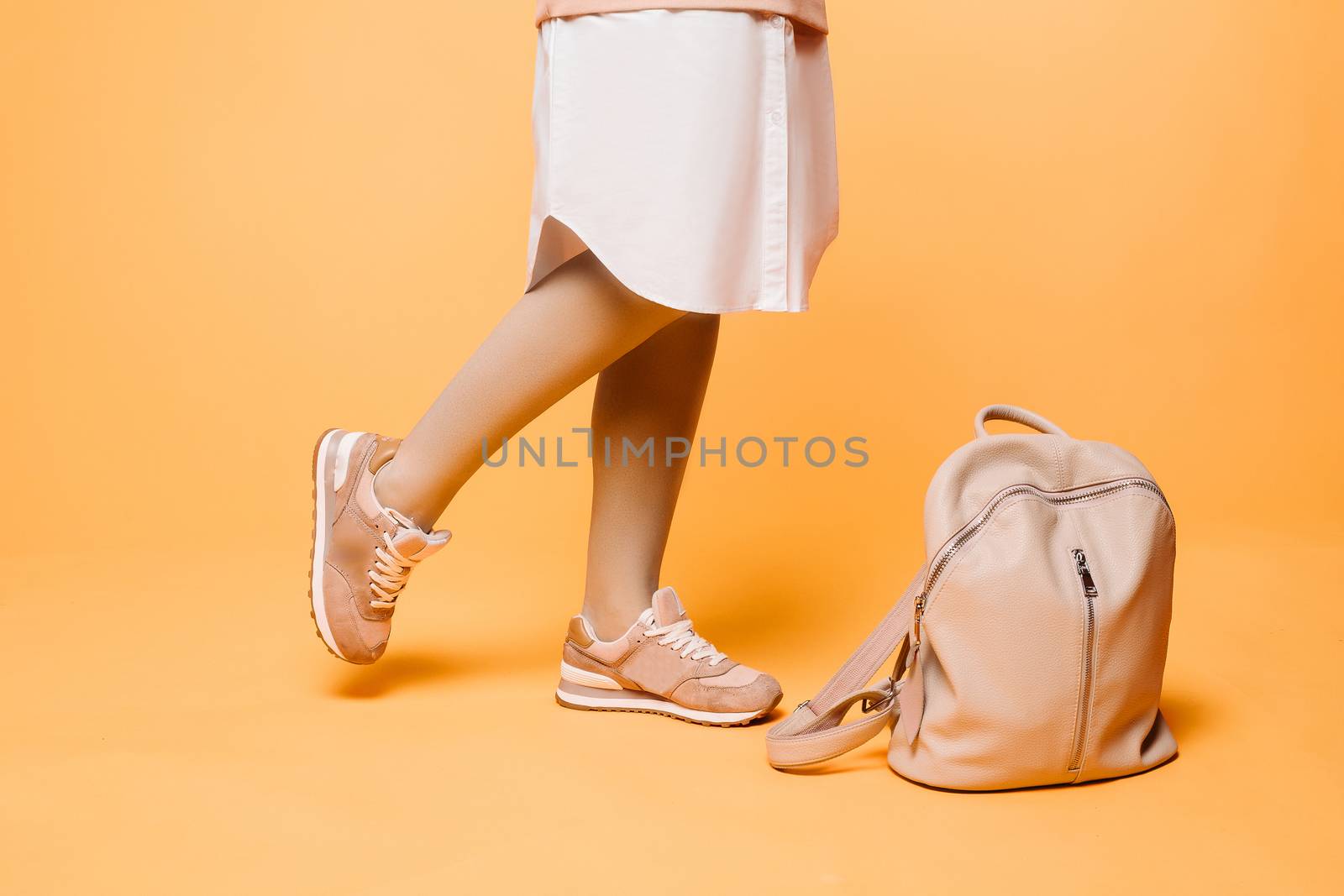 Crop of stylish incognito woman posing at studio in fashionable beige dress with elements of white, standing near pink leather bag on floor.Swag look of hipster woman against yellow studio background.