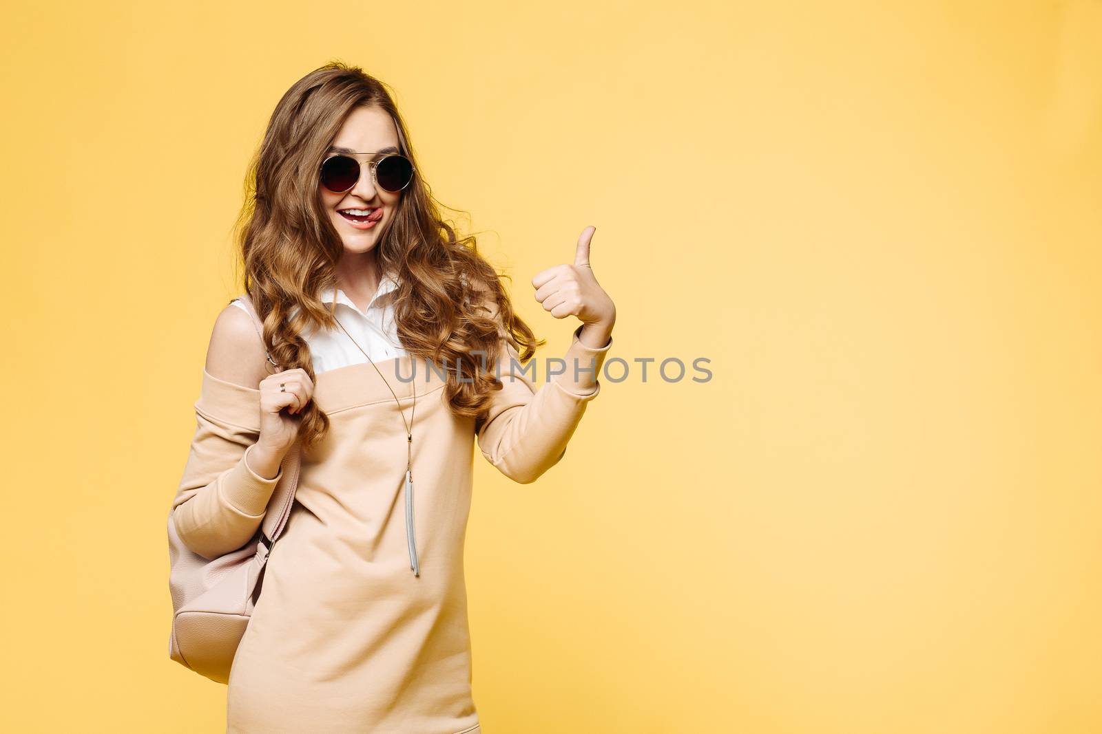 Beautiful fashionable brunette woman in sunglasses, posing at studio, rising up, holding leather bag over shoulder. Pretty and swag hipster girl with wavy hair, in beige dress after shopping.