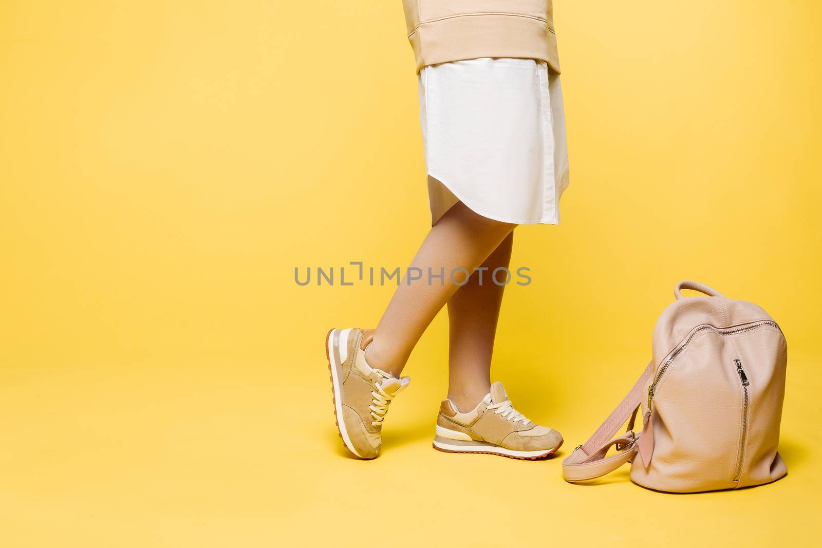 Crop of stylish incognito woman posing at studio in fashionable beige dress with elements of white, standing near pink leather bag on floor.Swag look of hipster woman against yellow studio background.