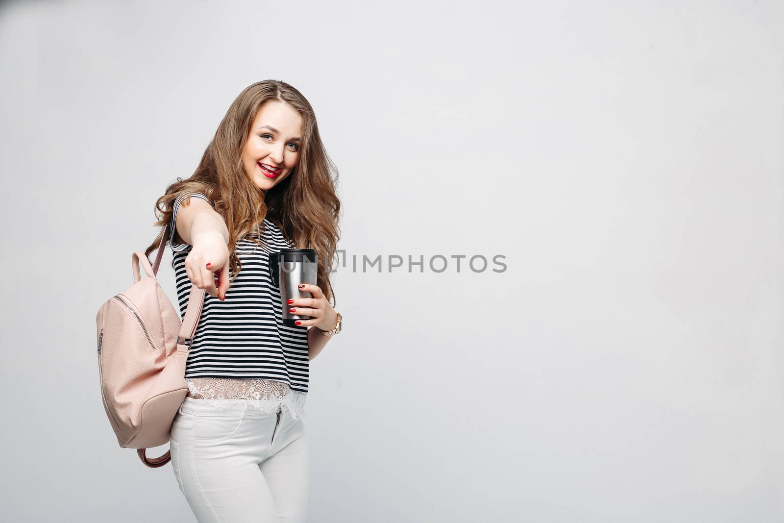 Pretty and stylish brunette girl with red lips, long wavy hair after beauty salon and shopping, posing at studio, holding thermos with warm coffee or tea. Sensuality fashionable woman looking down.