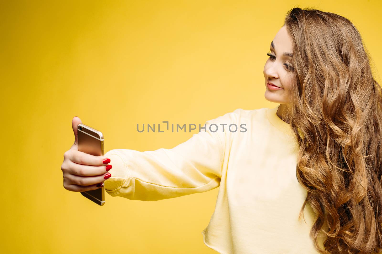 Stylish smiling beautiful brunette woman in yellow sweatshirt, posing and making self portrait at call phone. Fashionable girl with long hair,makeup, perfect manicure using call phone, taking photo.