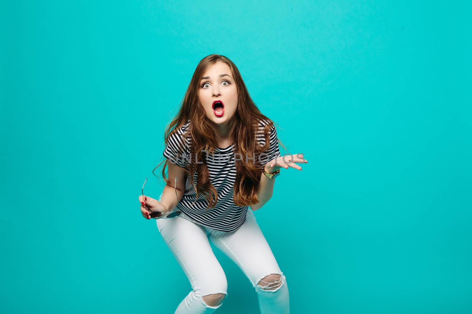 Emotionally brunette girl with long hair, stylish wearing in blouse and white jeans, gesturing by hands, shocking looking, screaming at camera. Woman surprised posing with opened mouth. Blue background.