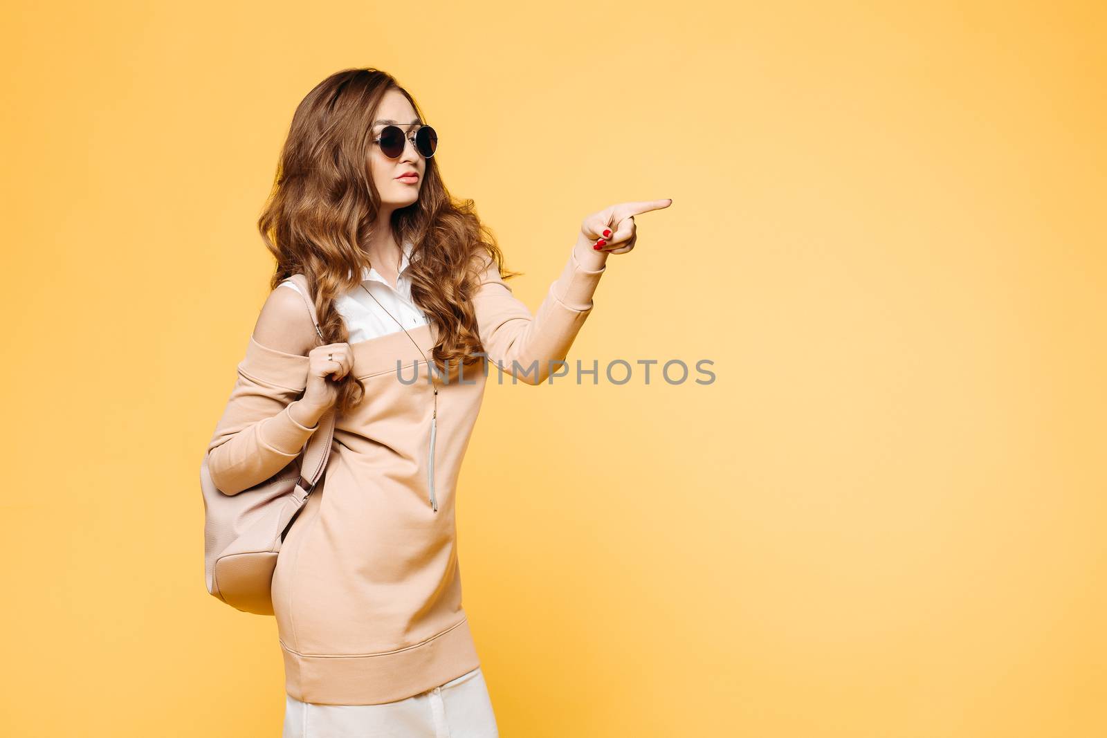 Beautiful fashionable brunette woman in sunglasses, posing at studio, rising up, holding leather bag over shoulder. Pretty and swag hipster girl with wavy hair, in beige dress after shopping.