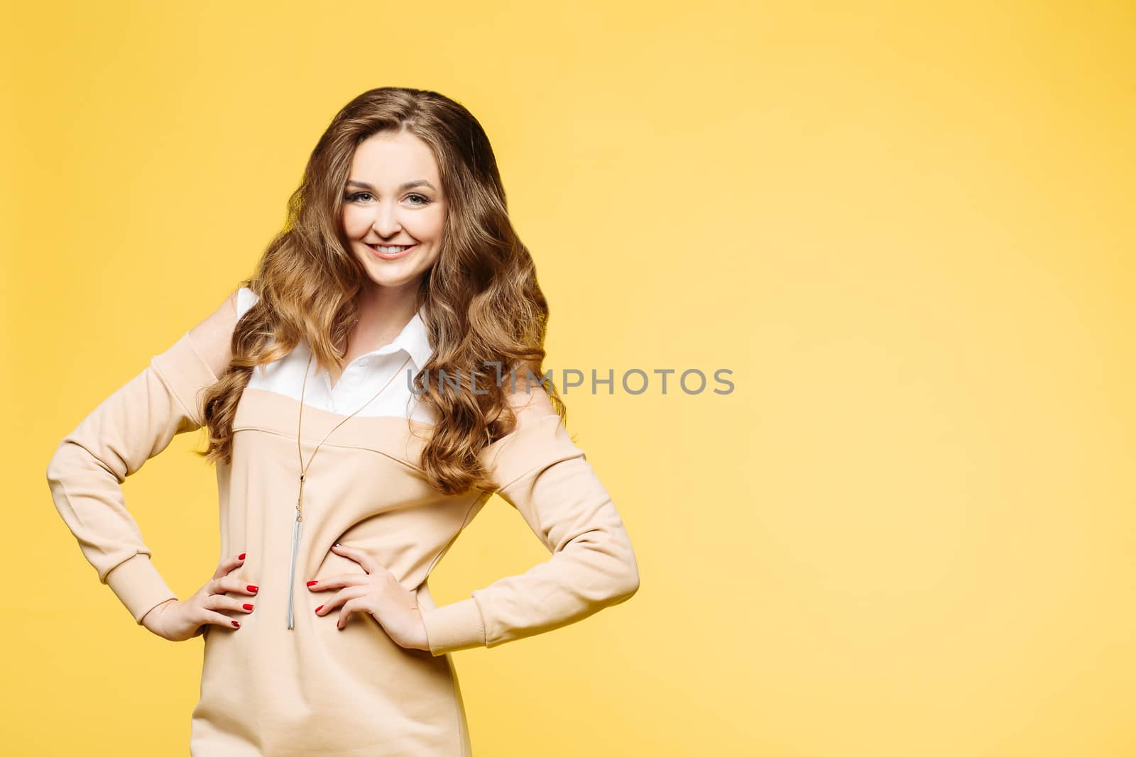 Fashionable beautiful brunette girl with wavy hair, perfect make up after beauty salon, wearing in white shirt and beige sweatshirt posing with hands on waist. Woman after shopping. Yellow background.