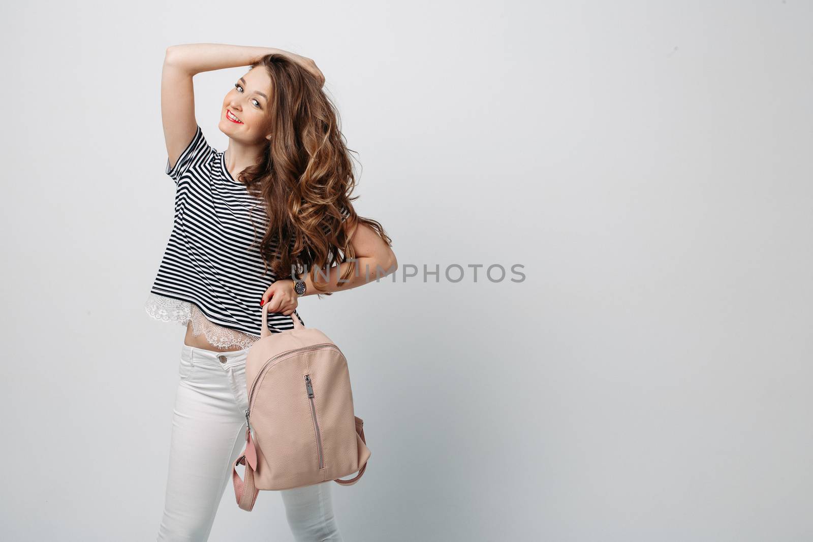 Fashionable and stylish woman with long wavy hair,red lips making duck face, surprised looking up. Girl wearing in blouse, white jeans, with leather bag, dreaming and touching face by finger. Fashion and advertising concept.