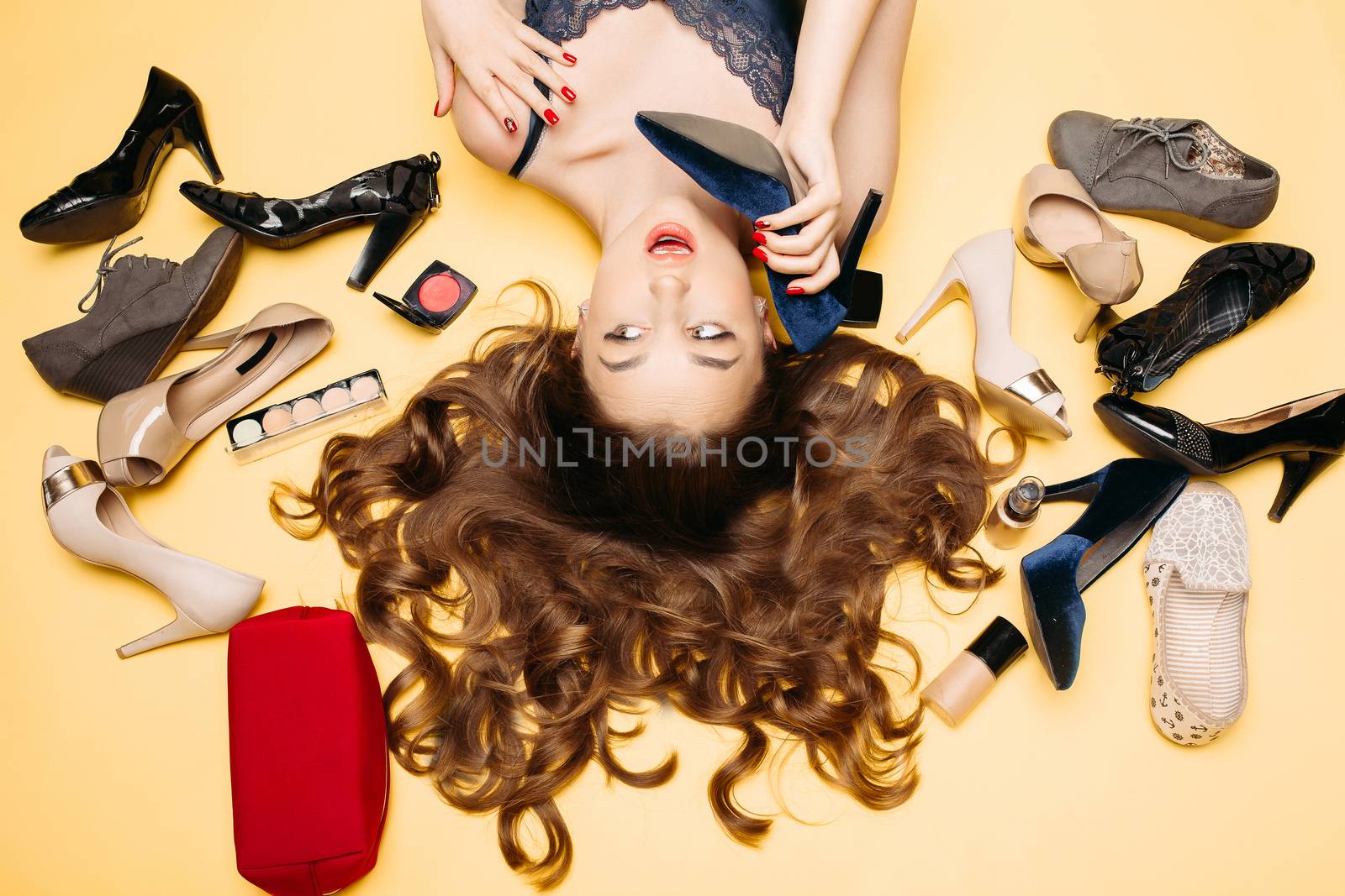 Fashionable brunette woman with long curly hair lying on yellow flor among many colorful shoes and laying, imitating conversation of like call phone. Beautiful girl emotionally posing, holding heel.