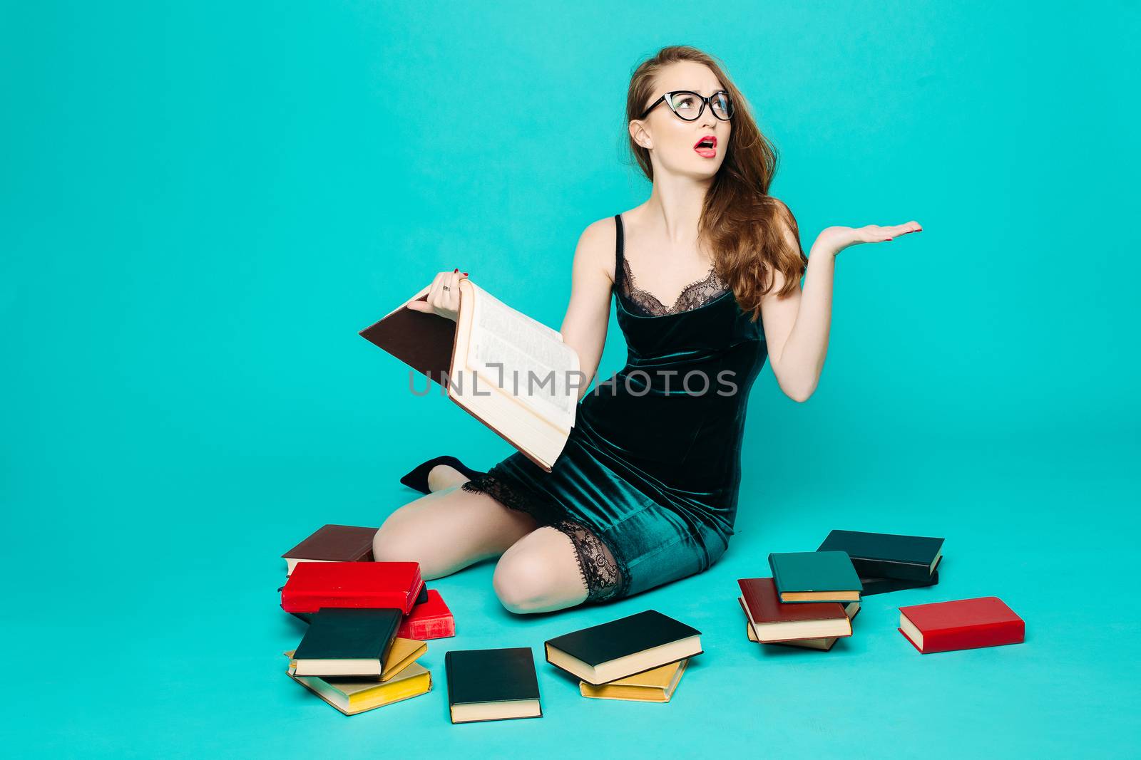 Sexy and beautiful female teacher in dress and eyeglasses, sitting on floor, posing and reading book, gesturing by hands, surprised looking up. Stylish pretty teacher or seductive female student.