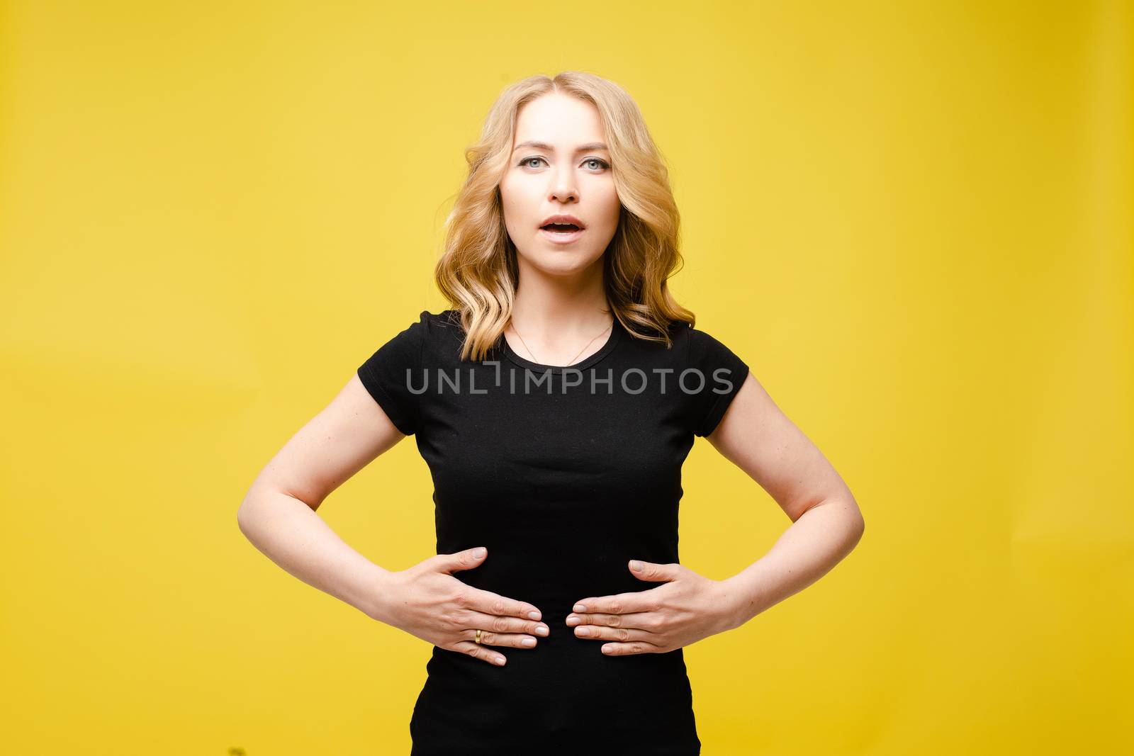 Stock photo of a blonde Caucasian woman in black t-shirt showing stomach ache. Woman holding her hands on her stomach demonstrating belly problems. Woman with open mouth demonstrating abdominal pain. Isolate on yellow.