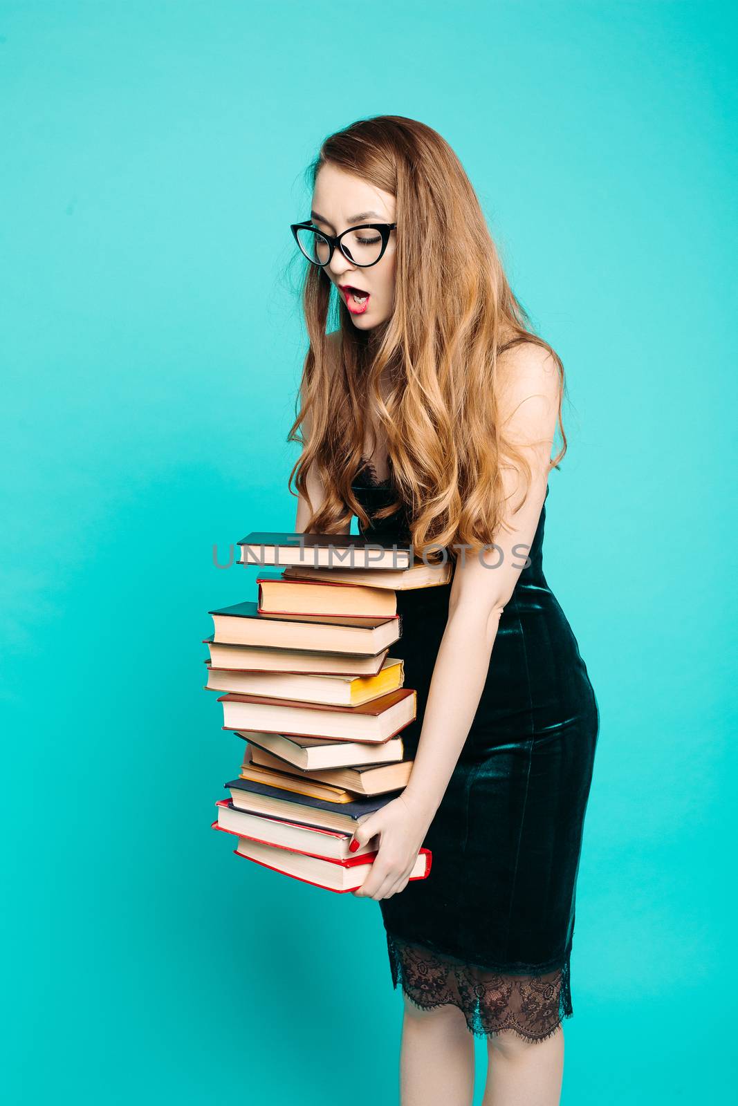 Portrait of emotionally shocked sexy teacher in dress with lace, eyeglasses, holding many books and surprised looking at camera. Young teacher or student screaming. Blue studio background.